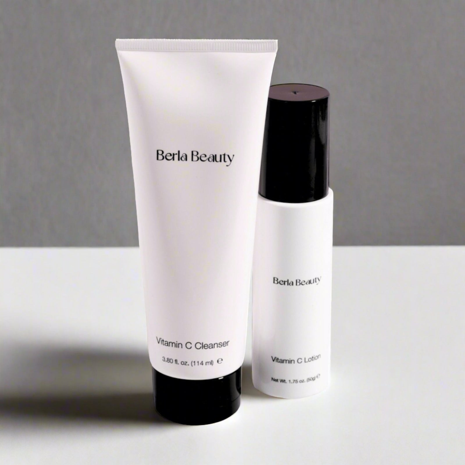 A white tube labeled "Berla Beauty Vitamin C Cleanser" stands next to a white bottle with a black cap labeled "Berla Beauty Vitamin C Lotion." This "Vitamin C Brightening Duo: Cleanser & Lotion Set" by Berla Beauty is designed for brightening your skin tone and includes a 3.80 fl. oz (114 ml) cleanser and a 1.75 fl. oz (50 ml) lotion.