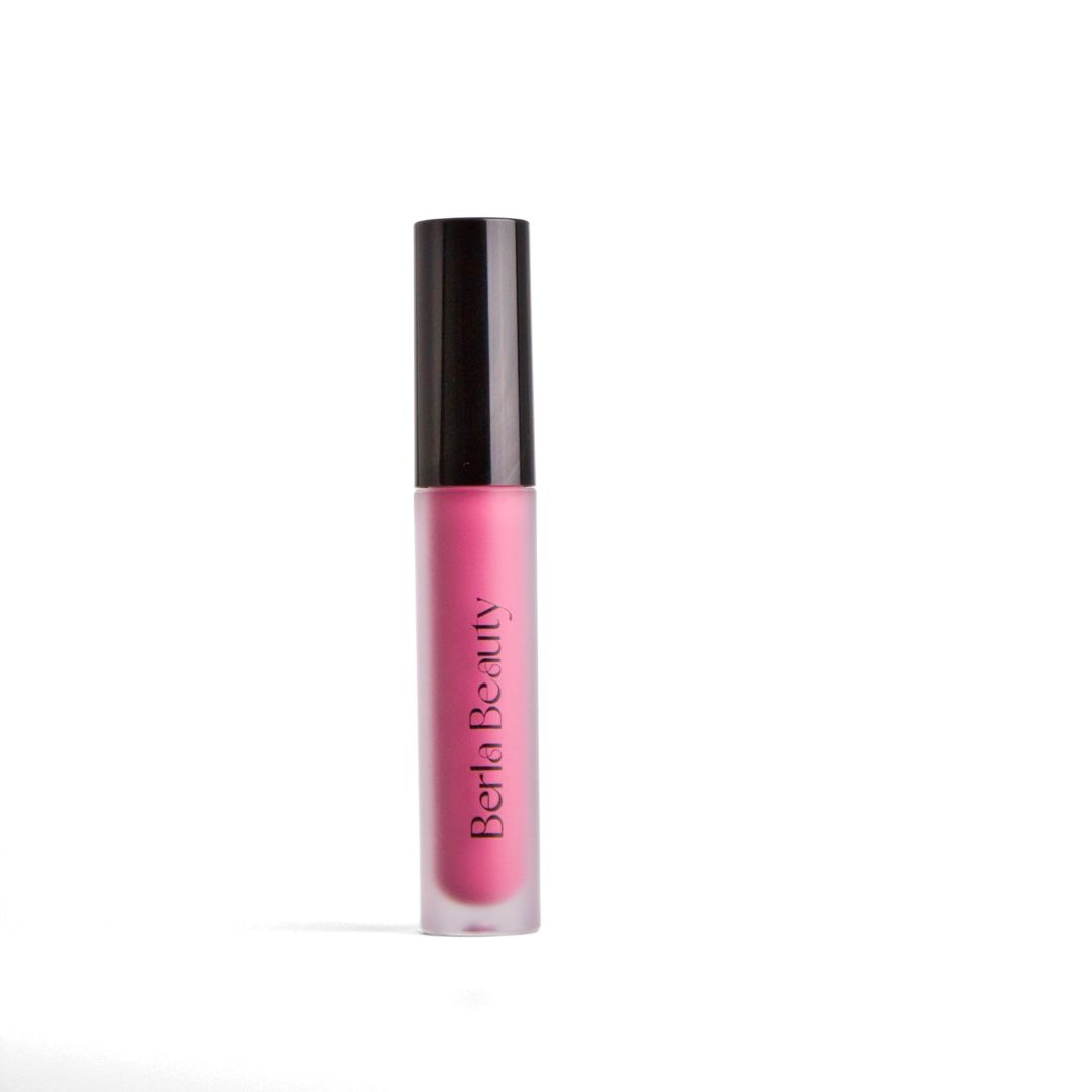 A tube of pink Brilliance Lip Gloss from Berla Beauty, which is cruelty-free and vegan, is open. The black cap with an attached applicator wand is placed beside it, showcasing the pink color on the applicator tip. The background is a soft beige color.