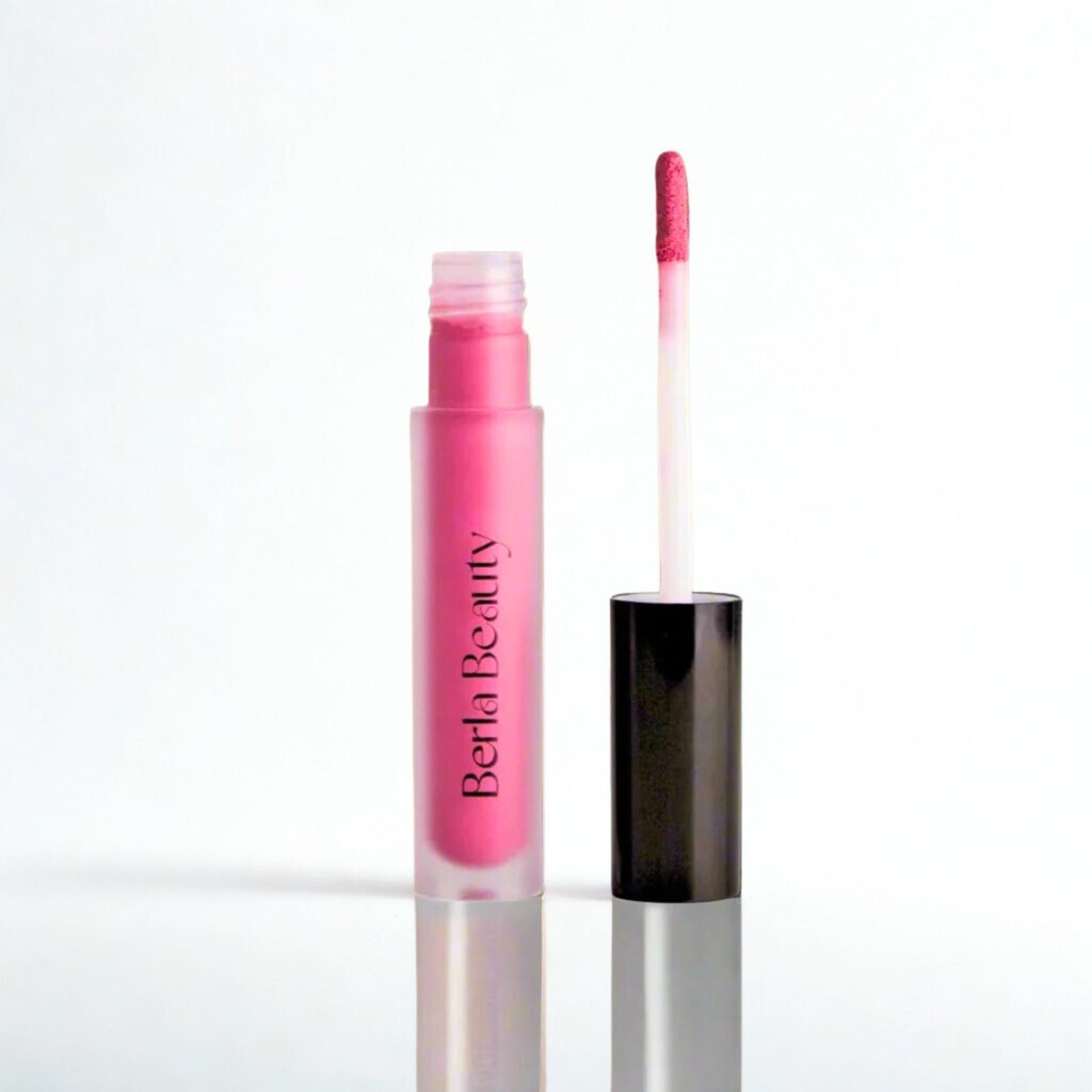 A tube of pink Brilliance Lip Gloss from Berla Beauty, which is cruelty-free and vegan, is open. The black cap with an attached applicator wand is placed beside it, showcasing the pink color on the applicator tip. The background is a soft beige color.