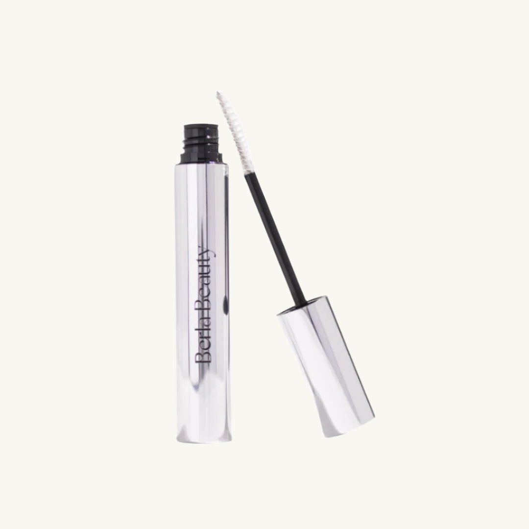 A sleek, metallic silver mascara tube with the brand name "Berla Beauty" printed vertically in black. The cap is off and resting beside the tube, revealing the wand with a white brush applicator designed for sculpted brows. The background is light beige. This product is the Vegan Clear Brow Gel - Long-Lasting Hold and Nourishing.