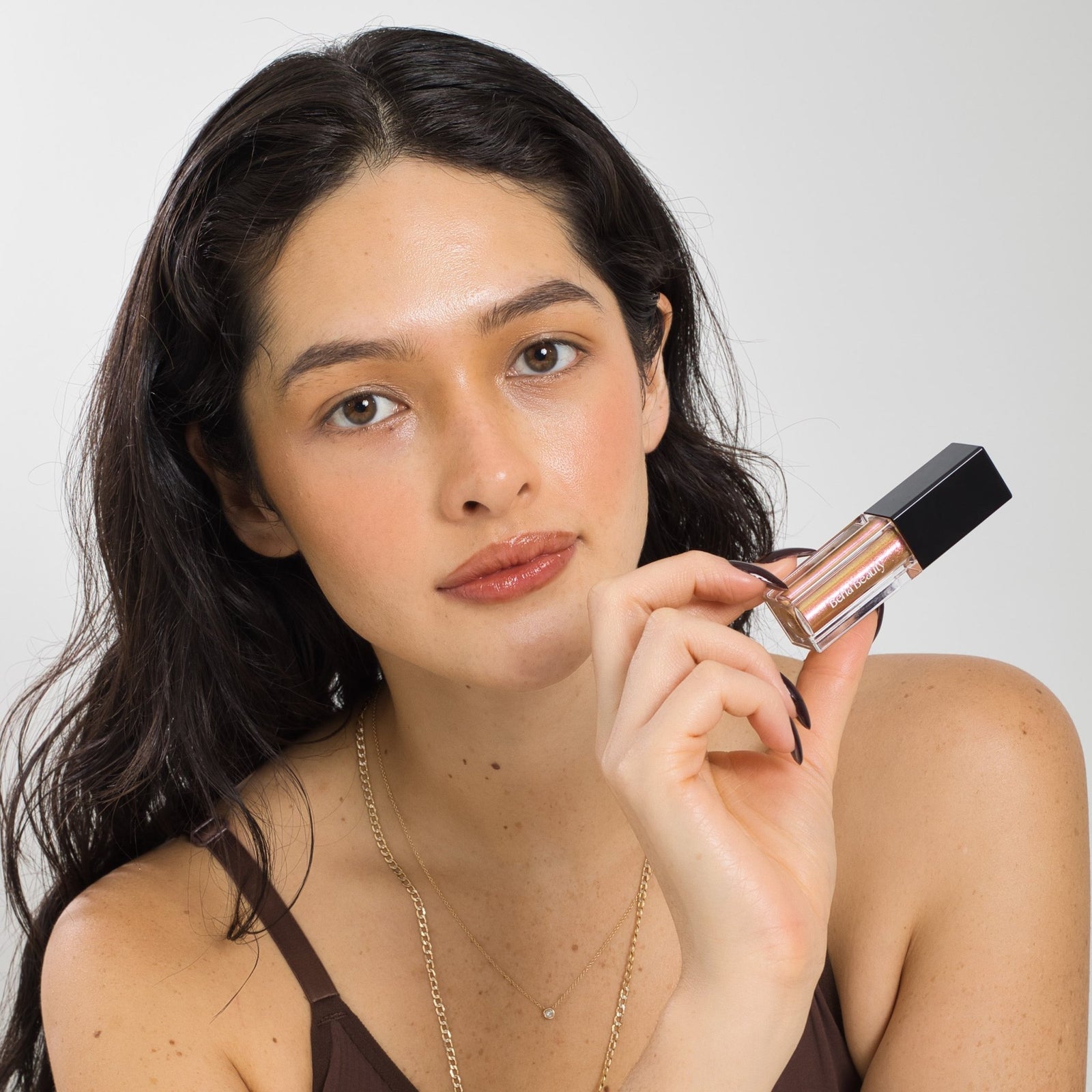 A rectangular liquid eyeshadow tube with a black cap is partially unscrewed, revealing a wand applicator. The eyeshadow boasts a long-lasting shimmer in an iridescent gold color and the product label reads "Berla Beauty." The background is a plain beige color.